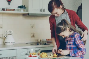 Mother helping daughter slice fruit at a kitchen counter while preparing a snack.